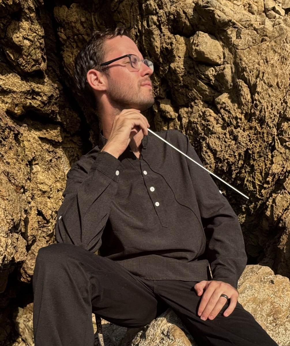 Jacob Boland, Founding Artistic Director of Resonaré Chamber Choir, seated outdoors holding a conductor's baton
